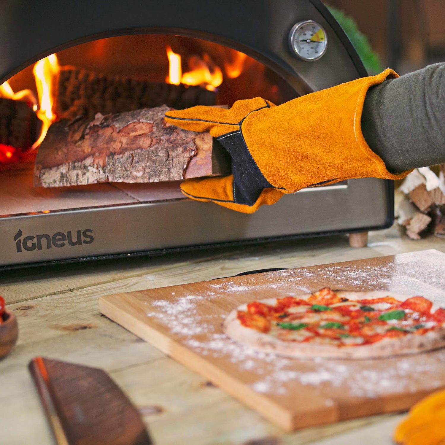 Igneus pizza oven heat resistant gloves inserting wood into stove with pizza in foreground